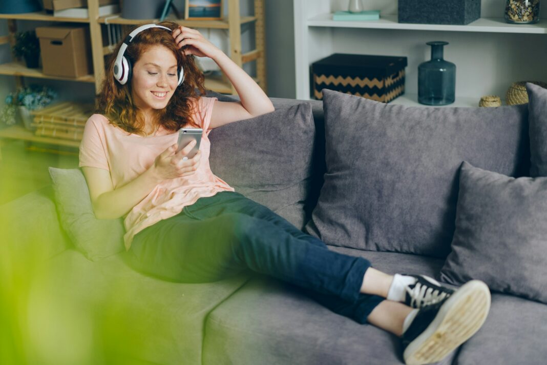 A woman sitting on her couch smiling at her phone with headphones on.