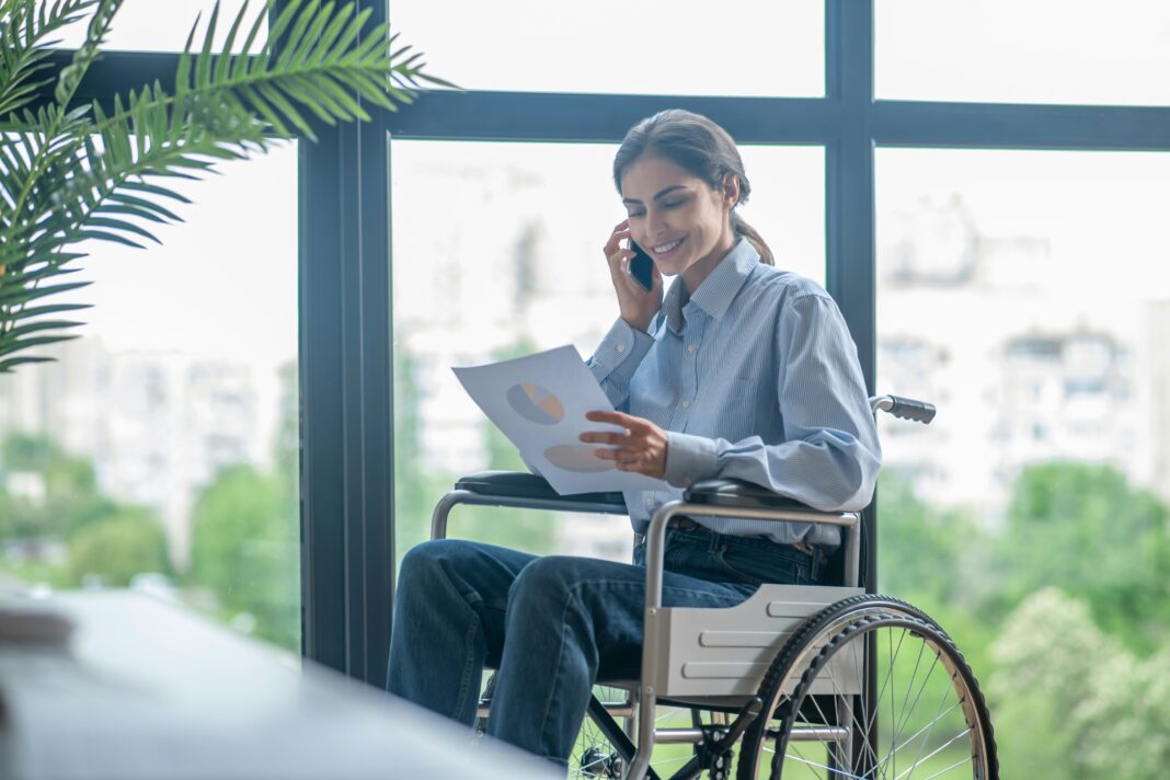 Photo of a woman sitting in a wheel chair, smiling and talking on the phone while looking at a piece of paper