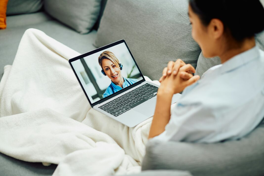 Woman sitting on her couch under a white blaket video calling someone on her laptop