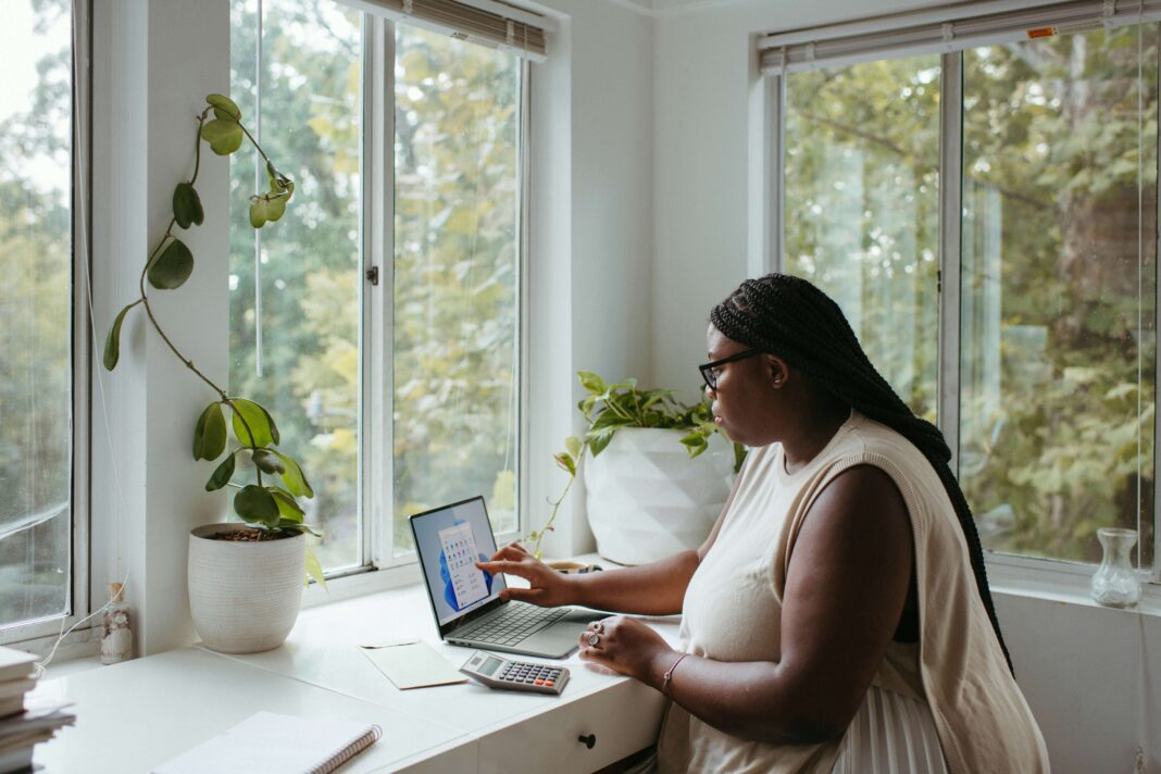 A woman sitting at her desk surrounded by windows and plants working on her laptop