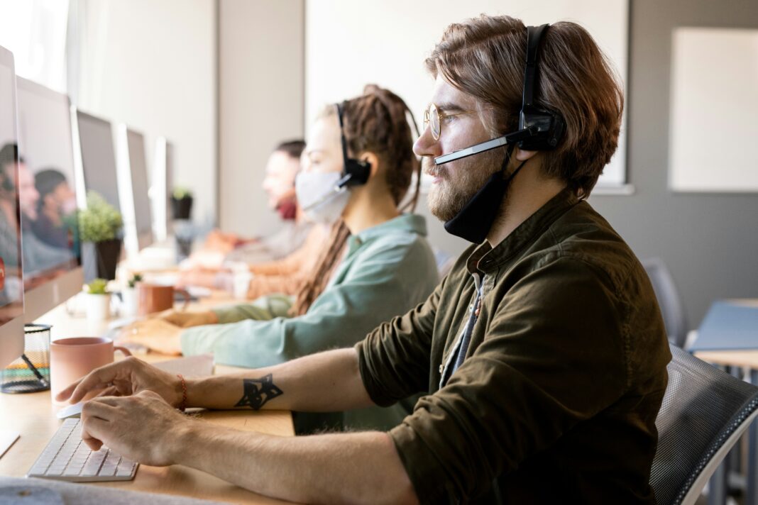 A man sitting at his desk next to a woman looking at a computer screen while wearing a headset