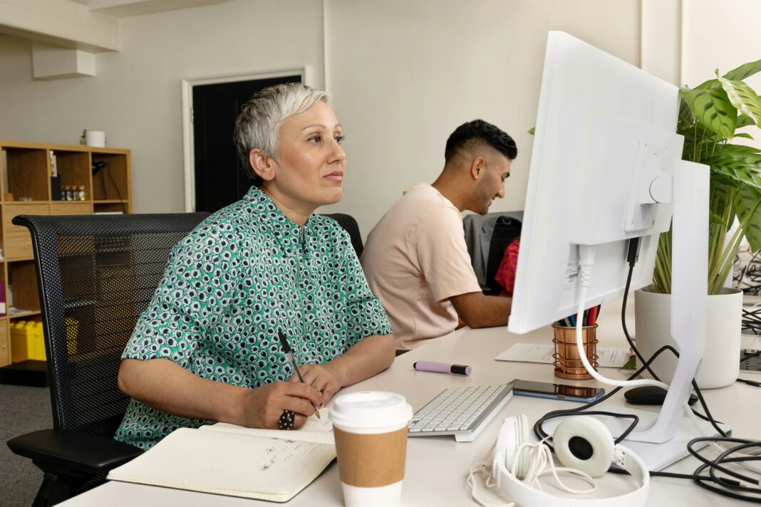 A woman sitting at her desk looking at her computer screen while writing in a notebook